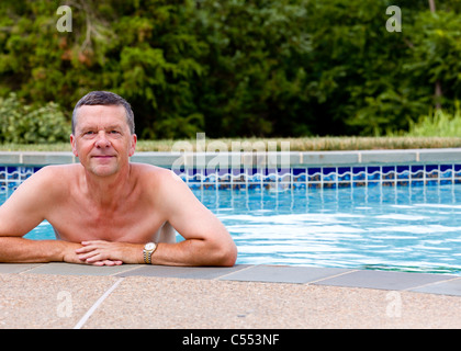 Senior male relaxing by the side of a modern swimming pool in back yard ...