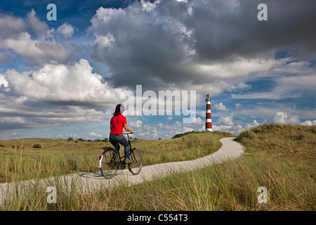 The Netherlands, Hollum, Ameland Island. Woman on bicycle on beach road. Lighthouse. Stock Photo