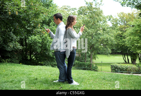 Young couple using their mobile phones in a park with backs against each other (posed by models) Stock Photo
