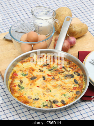 A freshly cooked Spanish omelet or tortilla de patates still in the pan, with essential ingredients behind Stock Photo