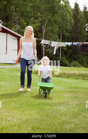 Full length of adorable child pushing a wheelbarrow in garden while walking with mother Stock Photo