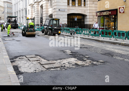 Street scene road works machines workmen & work in progress on resurfacing worn out tarmac road in City of London exposed manhole covers England UK Stock Photo