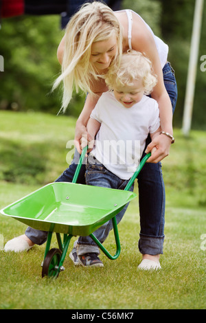 Young mother helping her baby boy in pushing wheelbarrow in garden Stock Photo