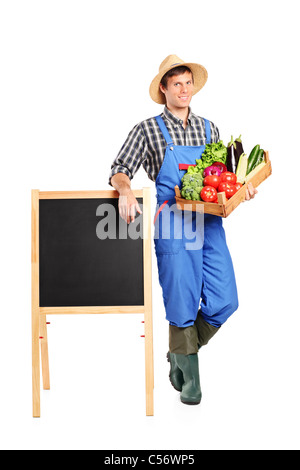 A farmer holding a crate full with vegetables and posing next to an empty board Stock Photo