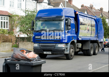 Black rubbish bin full to the brim waiting to be collected on a street in England as the bin lorry approaches in the background Stock Photo
