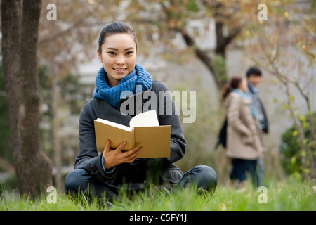 A young woman sitting on a grassy area reading a book, people in the background Stock Photo