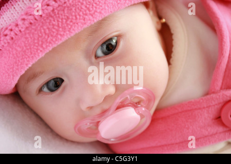 A cute baby girl with blue eyes and pink clothes lying on a crib with a pacifier in her mouth. Stock Photo