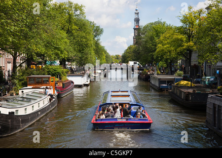 Tourists on a canal boat sailing on Prinsen Gracht Canal, Amsterdam, with the spire of Westerkerk Church in the distance.