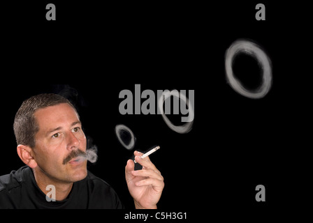 A man smoking a cigarette blows smoke rings against a black backdrop. Stock Photo