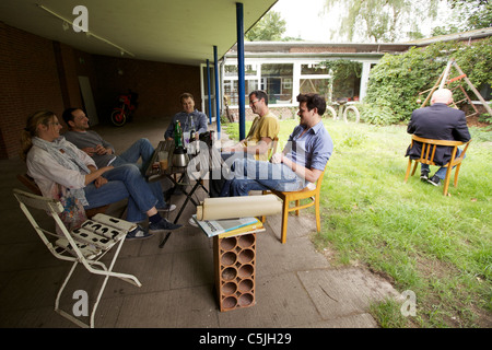 Members of the E1 artists' group sit under the covered porch of their atelier on Viktoriastraße in Grevenbroich, Germany, a motorcycle leaning against the facade in the background and another man sitting with his back to the camera at a separate table on the right. 23 July 2011. Stock Photo