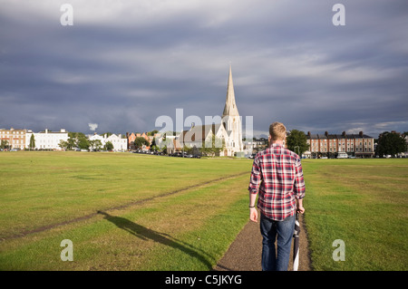 A young man walking across heath to village with All Saints parish church in evening sunlight. Blackheath, London, England, UK, Britain Stock Photo