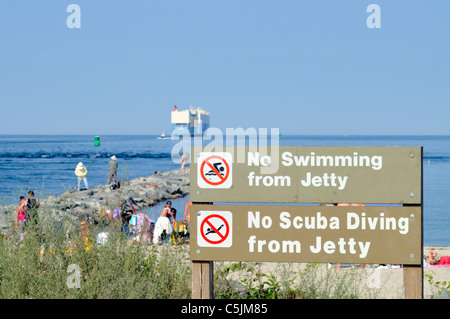 Warning signs about the jetty at the east end of the Cape Cod Canal with tourists fishing and tanning as a large cargo ship heads out to sea Stock Photo