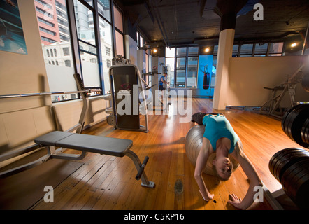 A gym in an old loft, with a young woman doing stretching exercises over a Swiss Ball, and a man working on a machine in the Stock Photo