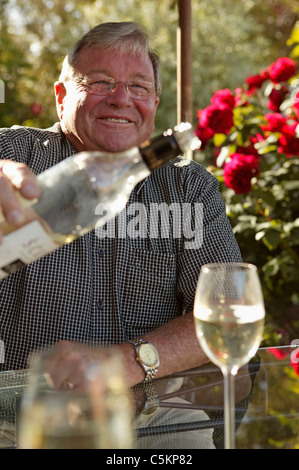 Man (60s) at home sitting at a table in his garden, smiling and pouring a glass of wine from a bottle, New Zealand Stock Photo