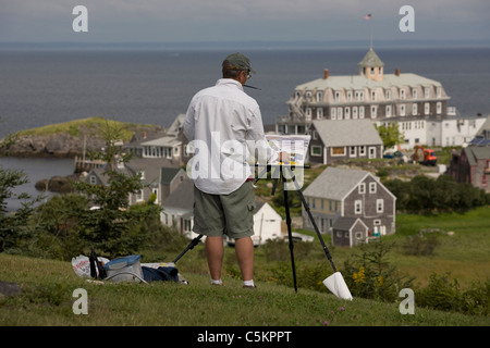 Monhegan Island, Maine -- Male artist paints using an easle atop a hillside overlooking Monhegan Island harbor and village and Stock Photo