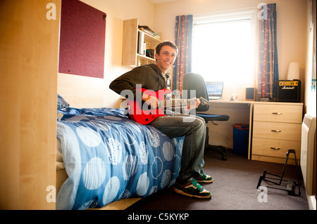 Young caucasian male student sitting on his bed in his room, in university halls of residence - playing a red guitar. Stock Photo