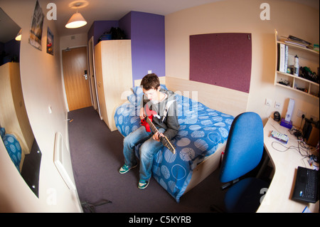 Young caucasian male student sitting on his bed in his room, in university halls of residence - playing a red guitar. Stock Photo