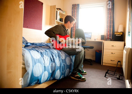Young caucasian male student sitting on his bed in his room, in university halls of residence - playing a red guitar. Stock Photo