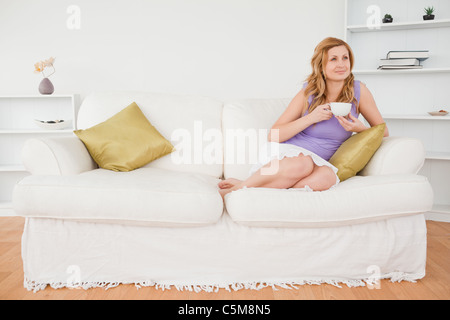 Pensive red-haired woman holding a cup of coffee and posing while sitting on a sofa Stock Photo