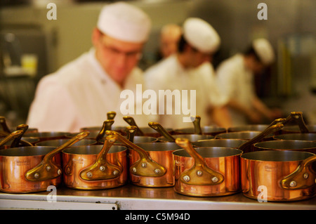 Copper pans in focus on a shelf in forground with chefs, out of focus working in the background in a busy  hotel kitchen. Stock Photo