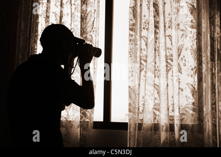 Man with binoculars looking out of window from behind net curtain: nosey neighbour, spy, undercover cop, pervert, stalker... concept Stock Photo
