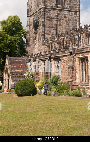 Holy Trinity Church, Skipton - 1 woman walking in sunny beautiful churchyard garden by south porch & stone clock tower - North Yorkshire, England, UK. Stock Photo