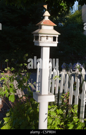 White bird house with metal roof and flag on top near sidewalk next to white picket fence. Stock Photo