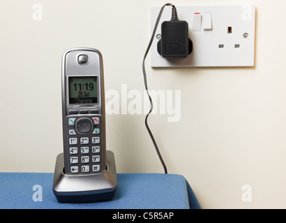 Cordless modern digital telephone handset charging on a phone charger base unit plugged into a wall socket power supply in a house. England UK Stock Photo