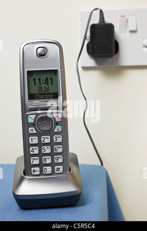 Cordless modern digital telephone handset charging on a phone charger base unit plugged into a wall socket. England UK Britain Stock Photo