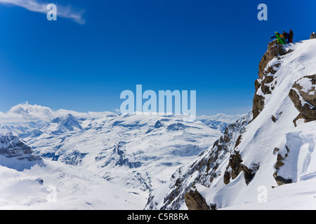 Three snowboarders standing on a snowy mountain look out over the summits and peaks below. Stock Photo