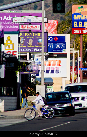 A lone bicyclist crosses the street as afternoon traffic passes retail advertising signs in Orange, CA. Stock Photo