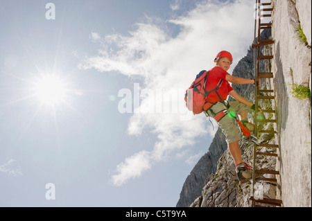 Germany, Garmisch-Partenkirchen, Alpspitz, Boy (12-13) climbing rock face on ladder, low angle view Stock Photo