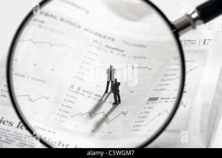 Two figurines on newspaper stock quotes with magnifying lens in foreground Stock Photo