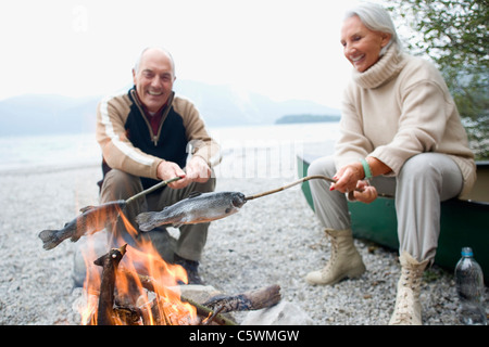 Germany, Bavaria, Walchensee, Senior couple sitting at campfire, grilling fish Stock Photo