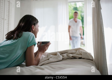 Germany, Hamburg, Woman lying on bed holding cup of tea, man in background Stock Photo