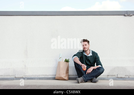 Germany, Berlin, Young man sitting in front of wall, holding an apple, portrait Stock Photo