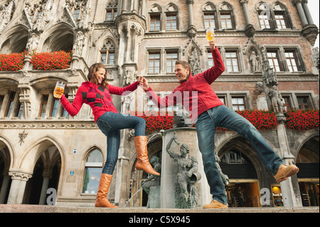Germany, Bavaria, Munich, Marienplatz, Couple balancing on fountain , holding beer mugs, smiling, portrait Stock Photo
