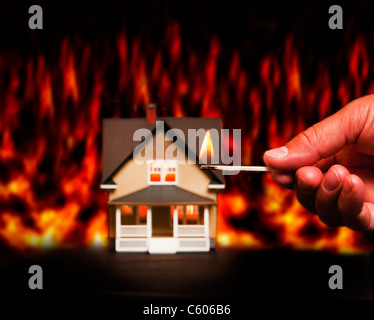 Close up of man's hand holding burning match with model house in fire in background Stock Photo