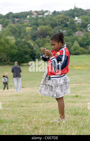 London Parliament Hill Hampstead Heath leisure pretty young black girl studiously making daisy or clover chain with Highgate Hill in background Stock Photo