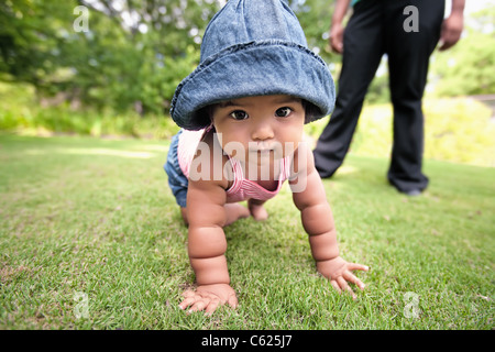 Baby girl crawling on grass, wearing a denim hat, with parent in background observing her chubby arms support her weight Stock Photo