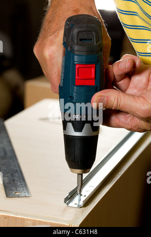 Screwing a screw into a drawer runner on side of wooden drawer using a battery drill Stock Photo