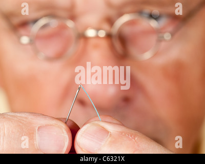 Macro image of a needle and cotton thread as senior man with glasses tries to thread the cotton