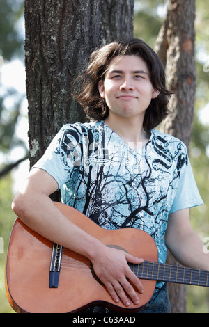 A Smiling Mexican Mixed Boy with Long Hair, Playing a guitar with Trees in the Background Stock Photo