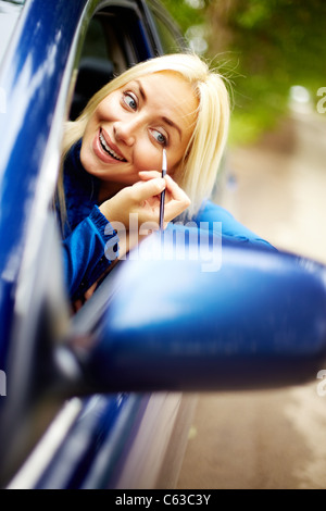 Photo of happy woman looking into car side mirror from car window while doing makeup