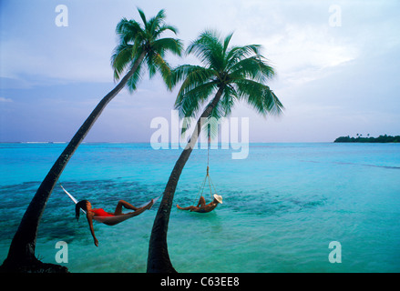 Couple hanging under separate palm trees over Indian Ocean waters on resort island of Meeru in the Maldives Stock Photo