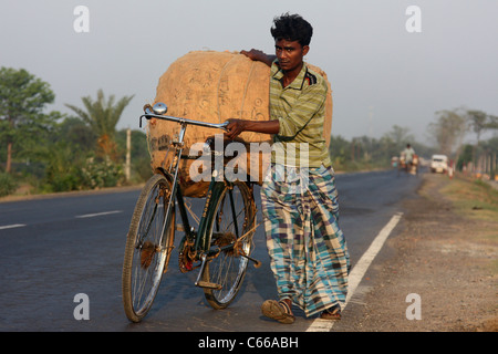 Young man struggles with heavy sack on bicycle up a busy highway at sunset in West Bengal India Stock Photo