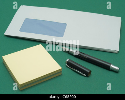 window envelopes on a green pad, one black pin and a block of adhesive labels in front of this Stock Photo