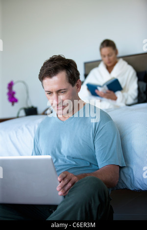 Mature man with laptop in bedroom and woman reading book in bed Stock Photo