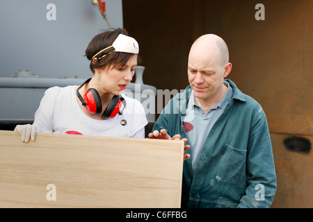 A man and a woman discussing the sanding work of a wooden door, wearing Personal Protective Equipment (PPE) for safety. Stock Photo