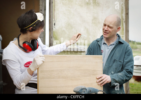 A man and a woman discussing the sanding work of a wooden door, wearing Personal Protective Equipment (PPE) for safety. Stock Photo